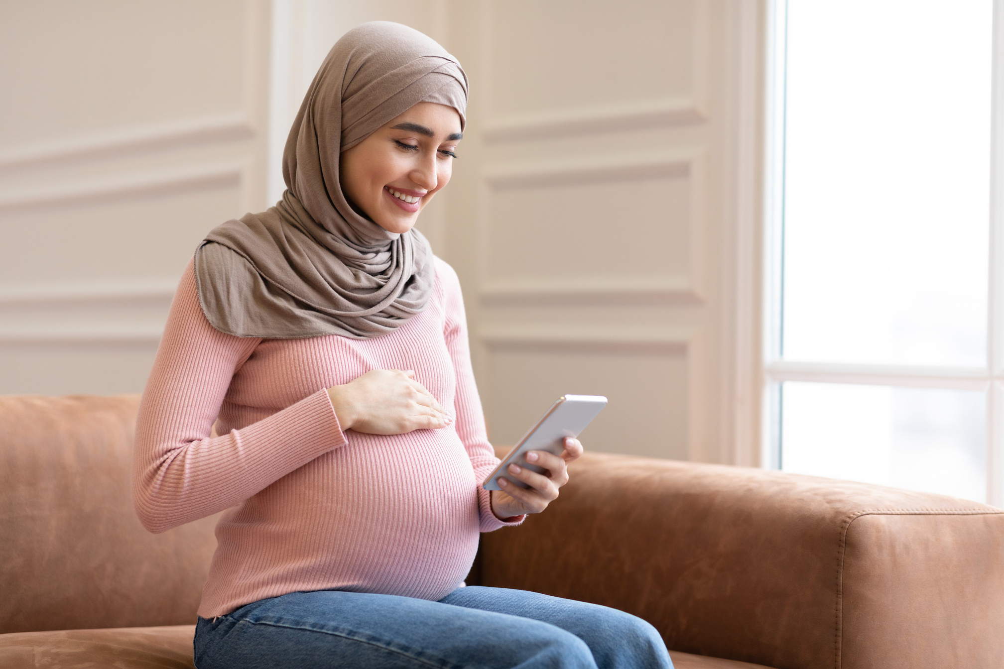 Happy Pregnant Muslim Woman Using Smartphone Sitting On Sofa Indoor